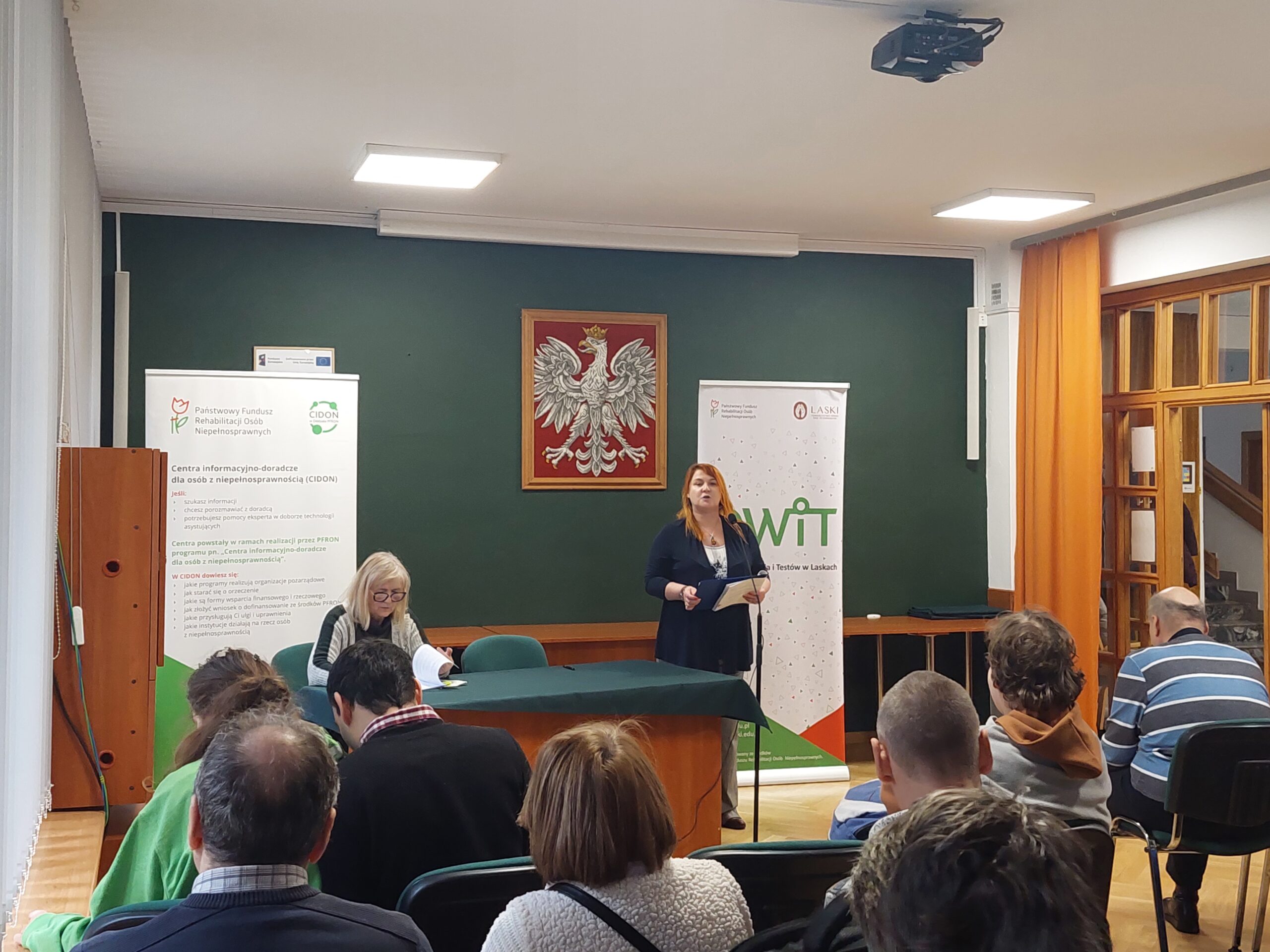 Przemawiająca do mikrofonu Pani prelegent z PFRON, oraz druga Pani prelegent siedząca przy stole