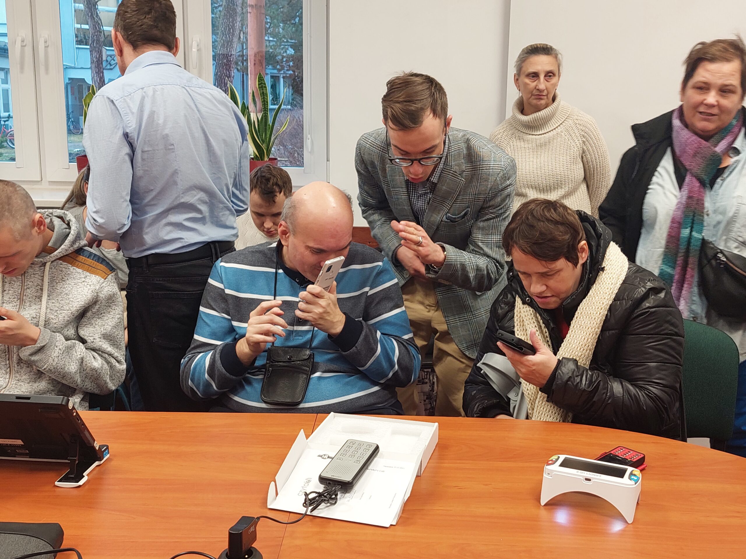 Uczestnicy oglądający różne sprzęty (telefony, lupy, powiększalniki) z pomocą pracowników OWiT